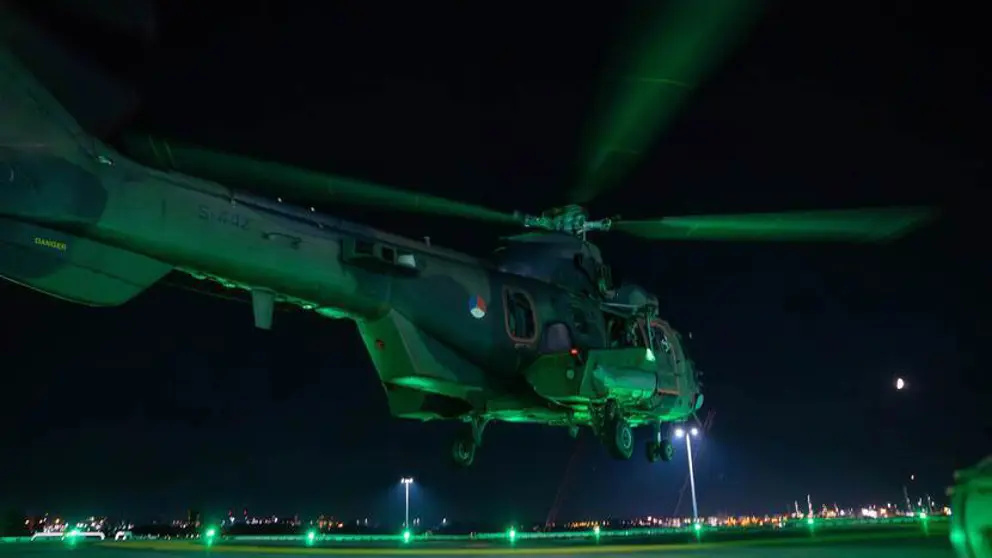 Groen aangelichte helikopter staat bijna aan de grond in het donker.