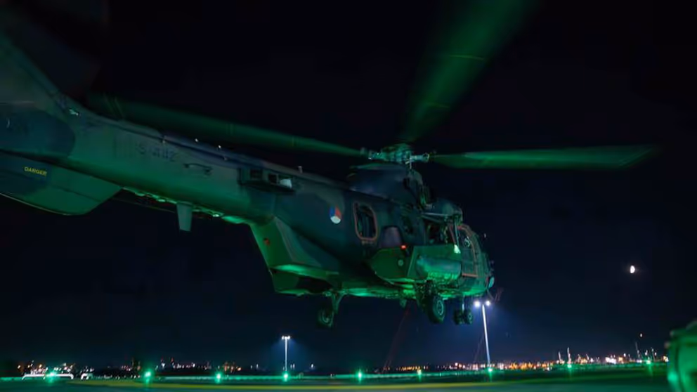 Groen aangelichte helikopter staat bijna aan de grond in het donker.