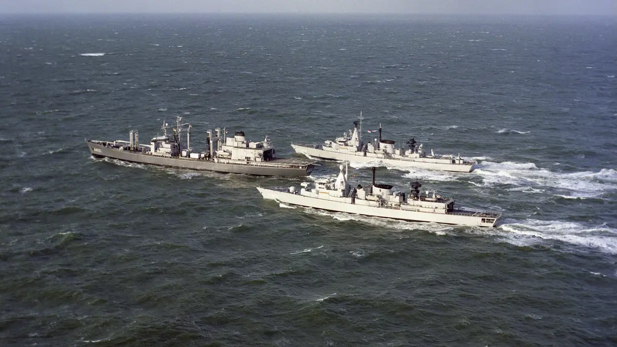 Drie marineschepen varen naast elkaar over een golvende, grijze zee onder een bewolkte lucht, met antennes en apparatuur op hun dekken.