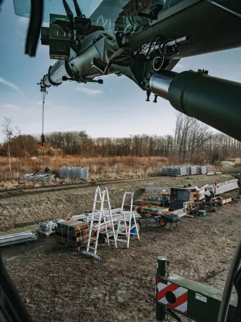 Het uitzicht vanuit een voertuig of machine met draaiende rotorbladen, gericht op een bouwplaats met materialen, ladders, kisten en stapels platen op een veld. 