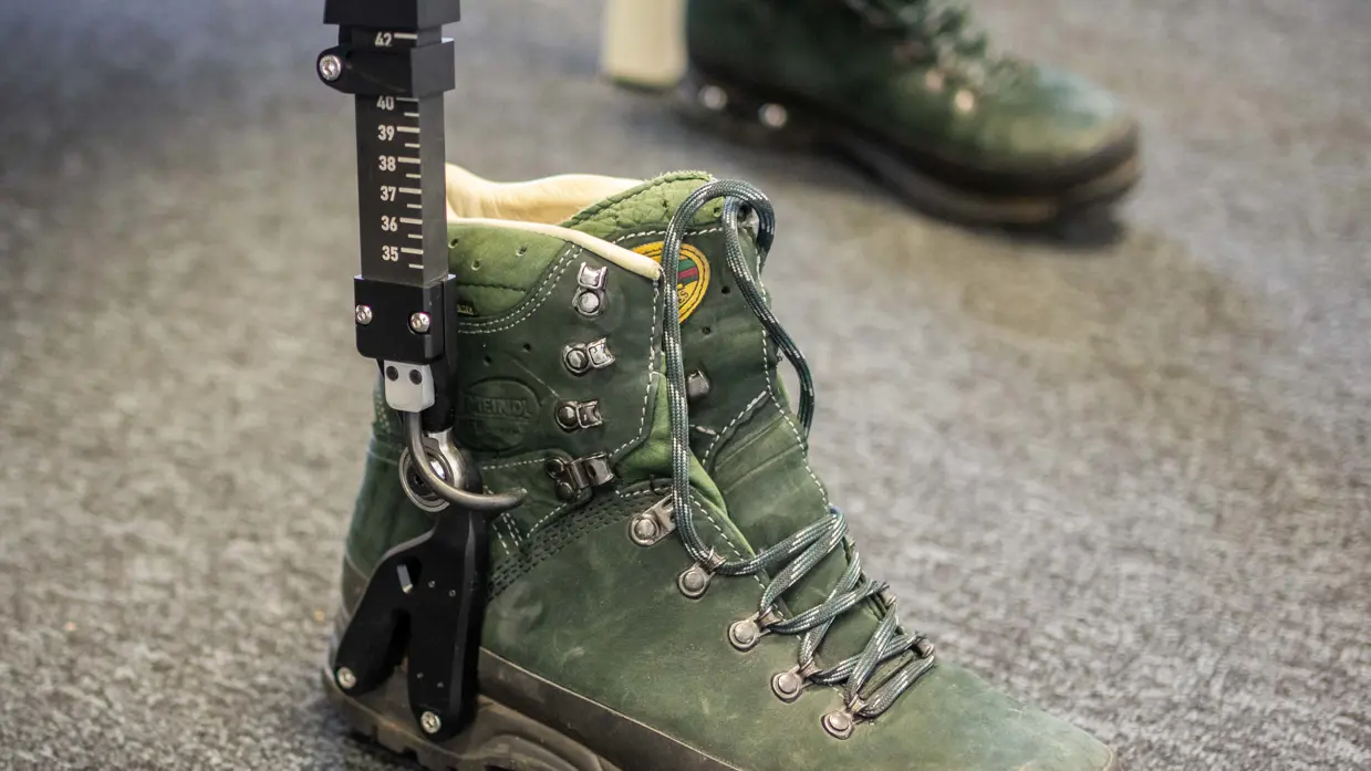 Een groene bergschoen met een zwarte metalen meetlat ernaast staat op een grijze vloer, tweede schoen op de achtergrond. 
