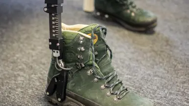 Een groene bergschoen met een zwarte metalen meetlat ernaast staat op een grijze vloer, tweede schoen op de achtergrond. 