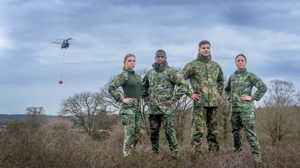 Twee vrouwelijke en twee mannelijke militairen poseren in de nieuwe kleding met het groene camouflagepatroon. Ze staan op de hei, op de achtergrond vliegt een Chinook met daaronder een oranje waterzak.