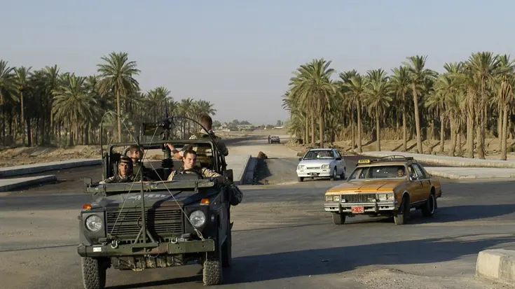 Militairen in een open jeep rijden over een weg met palmbomen; burgerauto's passeren op de achtergrond in een zonnige, droge omgeving. 