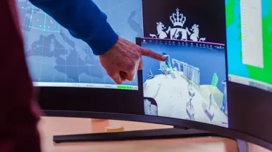Groep mensen staat rond stand op conferentievloer. Een man wijst op computerscherm in een stand op de conferentie en een man in blauwe trui op de rug gezien zit tussen 2 mannen met VR-bril op.