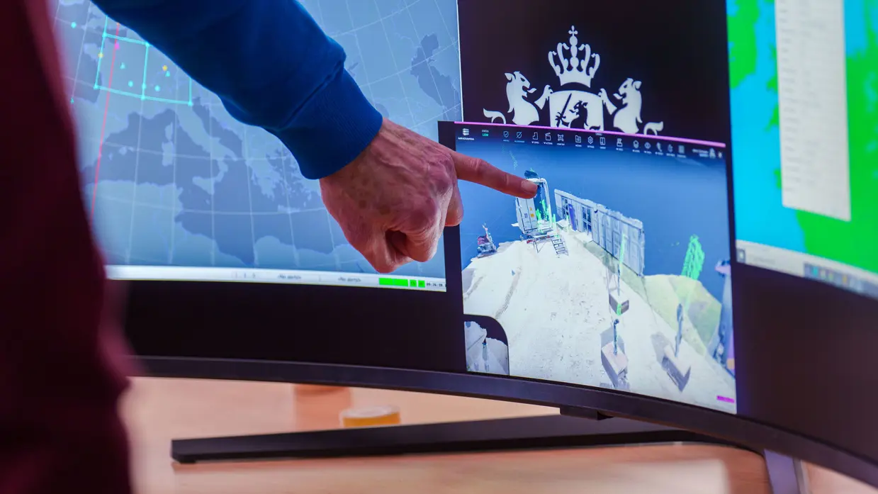 Groep mensen staat rond stand op conferentievloer. Een man wijst op computerscherm in een stand op de conferentie en een man in blauwe trui op de rug gezien zit tussen 2 mannen met VR-bril op.