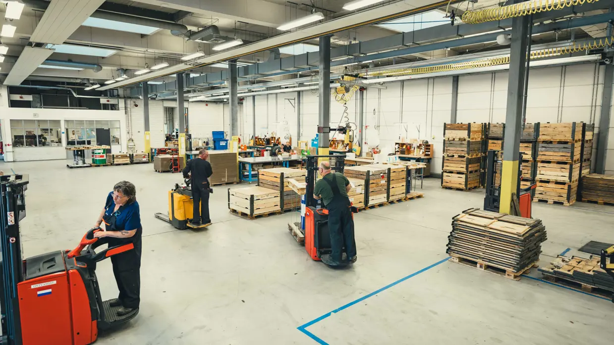 Overzicht van de grijze loods. Er zijn diverse lange tafels, boxpallets en heftrucks te zien. 