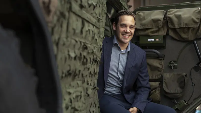 Een lachende man in blauw pak zit op een militair voertuig. 