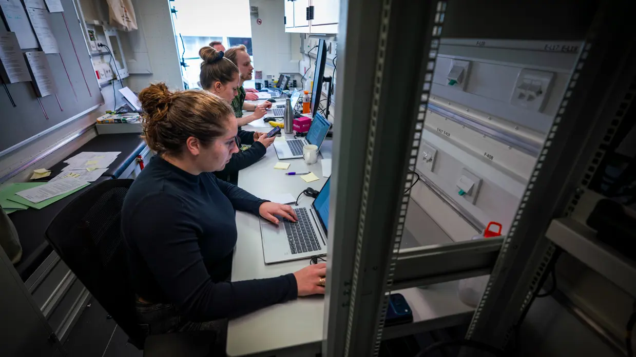 Twee vrouwen en twee mannen, waarvan één in uniform, zitten achter computers in een conrtainer.