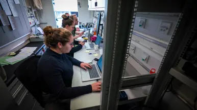 Twee vrouwen en twee mannen, waarvan één in uniform, zitten achter computers in een conrtainer.
