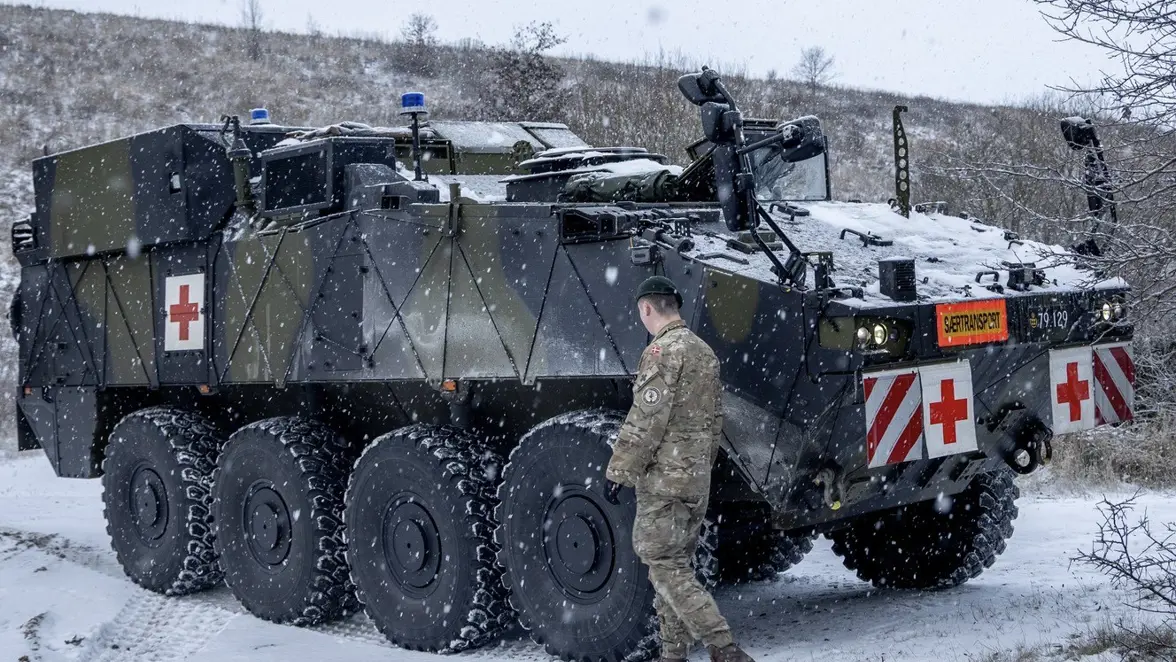 Een militair in winterkleding loopt naast een groot, zwaar pantservoertuig met rode kruizen en sneeuw op de weg en een ander voertuig, tijdens een sneeuwbui. 