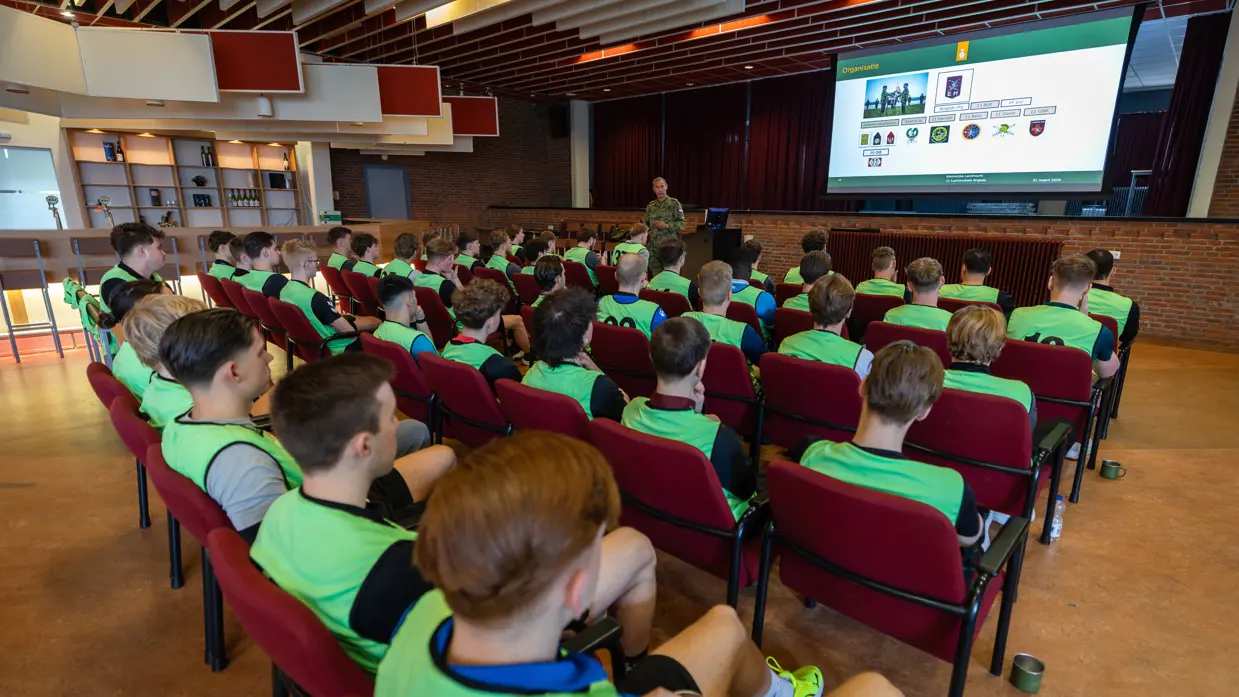 Een groep jongeren in gele vesten luistert in een zaal naar een militair die een presentatie geeft over organisatie en verschillende logo’s op een groot scherm.
