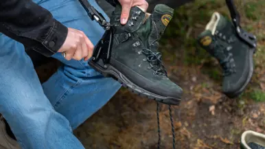 Iemand trekt een groene bergschoen aan met een zwarte beugel, buiten op een bosbodem met bladeren.