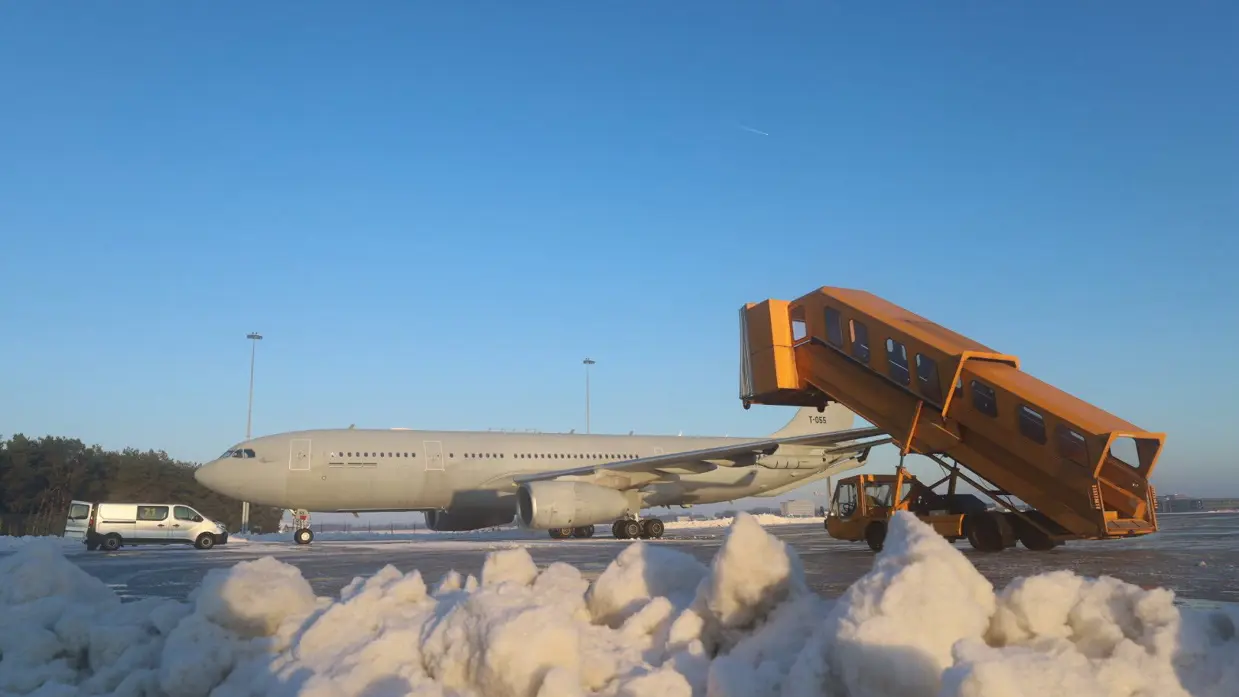 Links: drie gele sneeuwruimvoertuigen werken op een besneeuwde startbaan van een luchthaven, met een grijze lucht en sneeuw aan weerszijden. Rechts: Een Airbus 330MRTT staat op de besneeuwde luchthaven, met een oranje trap die naar de deur rijdt en sneeuwhopen op de voorgrond.