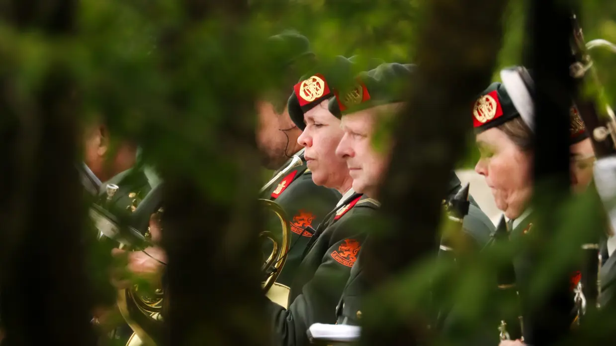 Een groene, wazige achtergrond met muzikanten in donkergroene uniformen en rode baretten die instrumenten bespelen. 