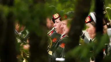 Een groene, wazige achtergrond met muzikanten in donkergroene uniformen en rode baretten die instrumenten bespelen. 