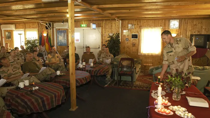 Militairen in uniform zitten in een houten ruimte; één man steekt kaarsen aan op een tafel met bloemen, boeken en theelichtjes. 