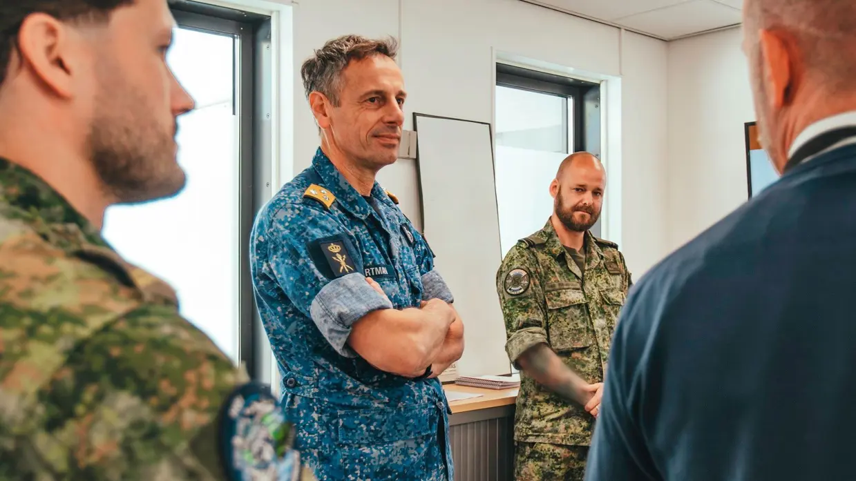 Vice-admiraal Jan Willem Hartman, in blauw gevechtstenue, staat tussen twee militairen in groen uniform. Hij luistert naar een derde persoon in net pak.