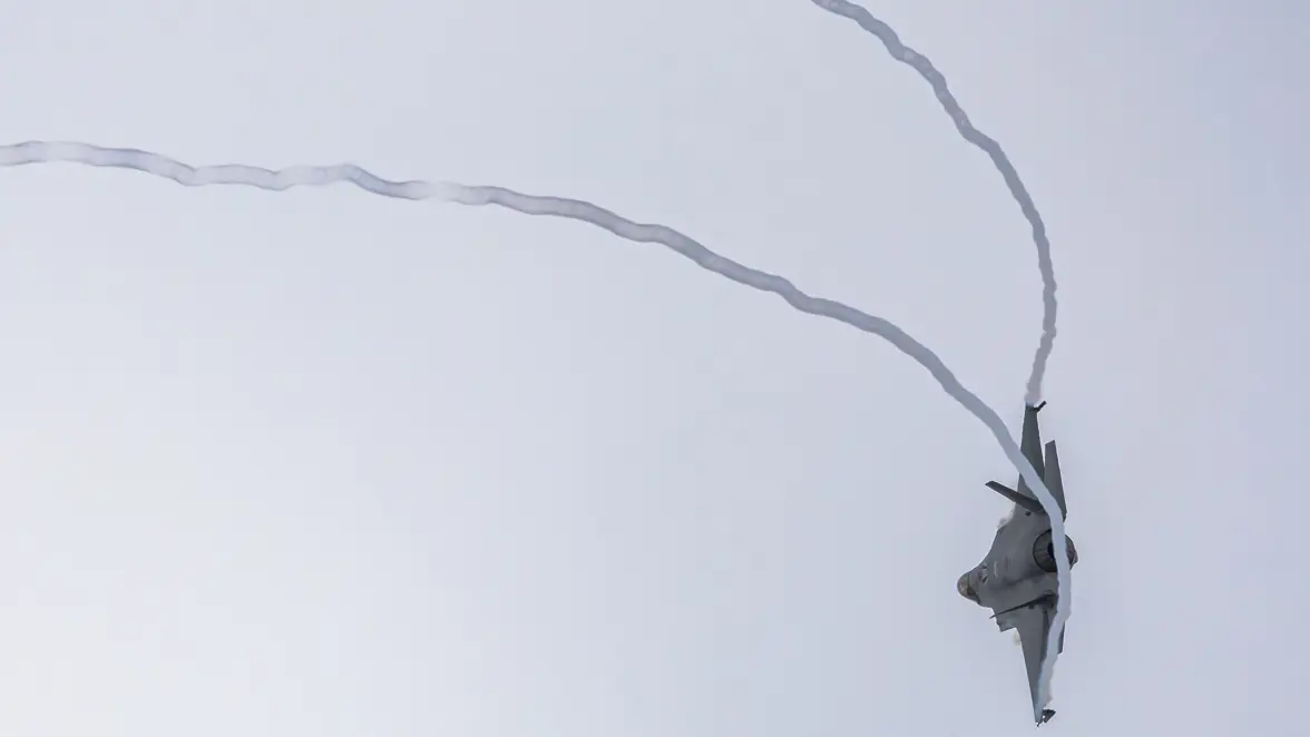 Een straaljager vliegt ondersteboven hoog in de lucht en trekt een witte rooklijn achter zich aan, tegen een lichte, heldere hemel.    
