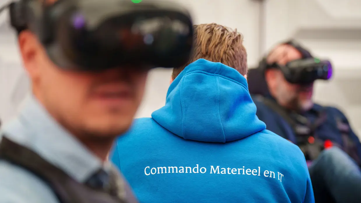 Groep mensen staat rond stand op conferentievloer. Een man wijst op computerscherm in een stand op de conferentie en een man in blauwe trui op de rug gezien zit tussen 2 mannen met VR-bril op.