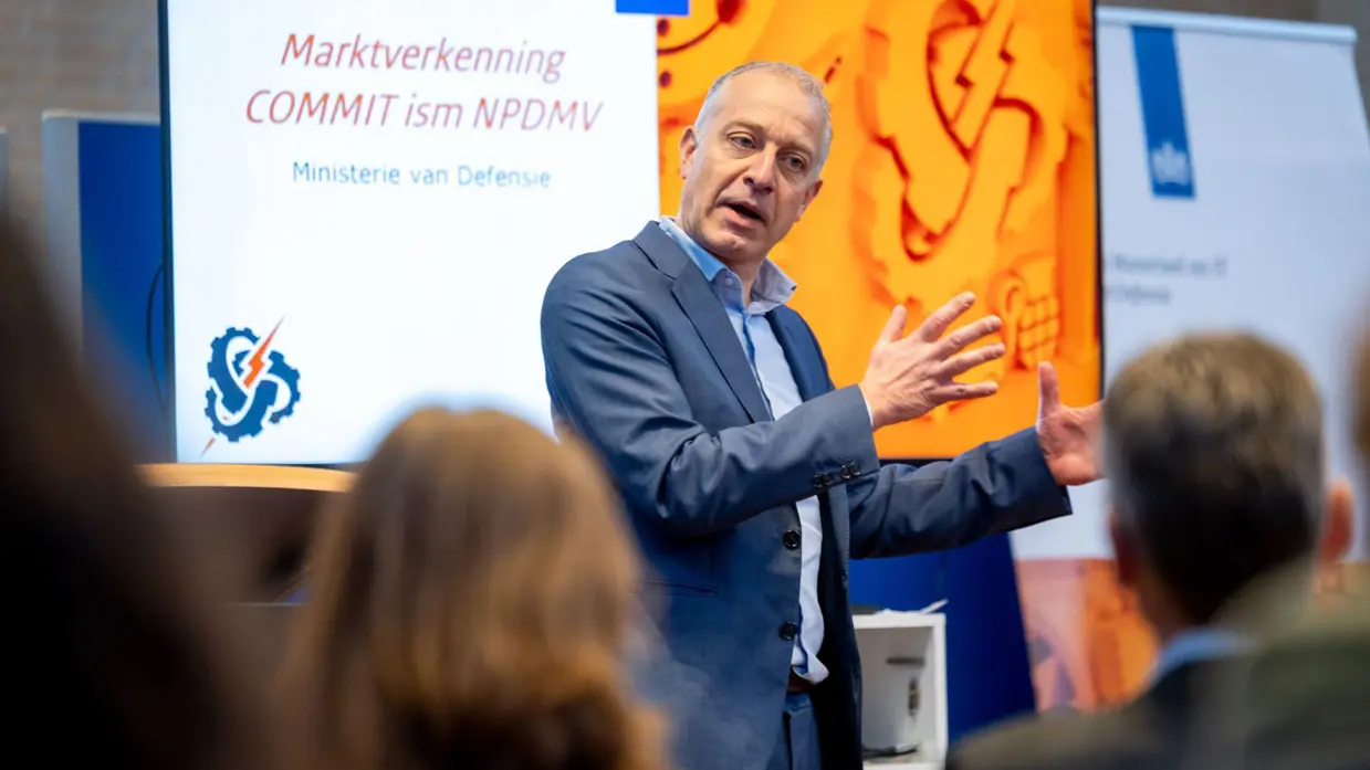 Een man in pak spreekt voor een groep mensen en maakt daarbij gebruik van zijn handen. 