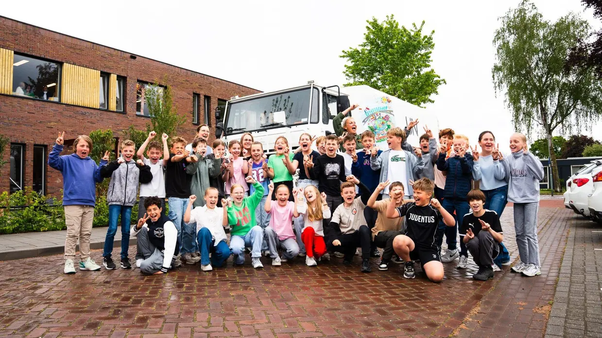 +alttekst: Groep blije kinderen en volwassenen poseren voor een witte bus met kleurrijke tekeningen, bij een rood bakstenen gebouw. 