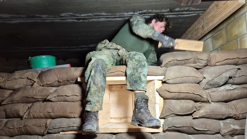 Een persoon in camouflagekledij leunt tegen zandzakken, met zijn voeten op een houten kist, in een schuilplaats.