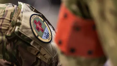 Links: Een militair werkt geconcentreerd achter een bureau met computerschermen. Op de schermen zijn kaarten te zien, terwijl een collega op de achtergrond ook bezig is. Rechts: Een close-up van een militair uniform met een rond embleem ‘Casualty Move 2026’ en een rood kruis. Op de achtergrond is wazig een persoon in oranje zichtbaar.