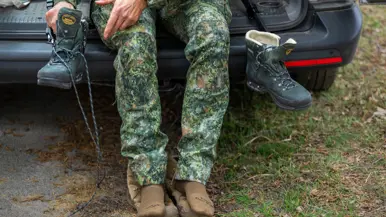 Iemand in camouflagebroek zit op een auto met bruine sokken en houdt een groene bergschoen vast.