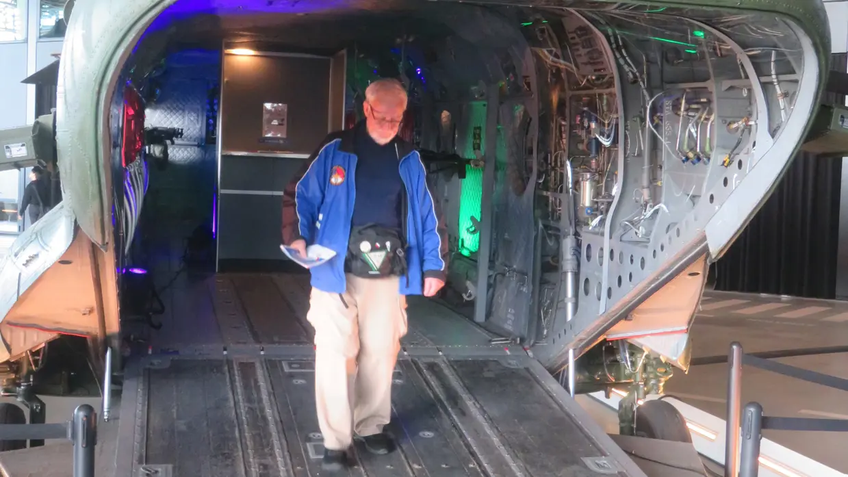 Inkijk in een grote, openstaande militaire helikopter; binnenin zie je stoelen, apparatuur en knipperende groene lichten, terwijl iemand binnen staat.