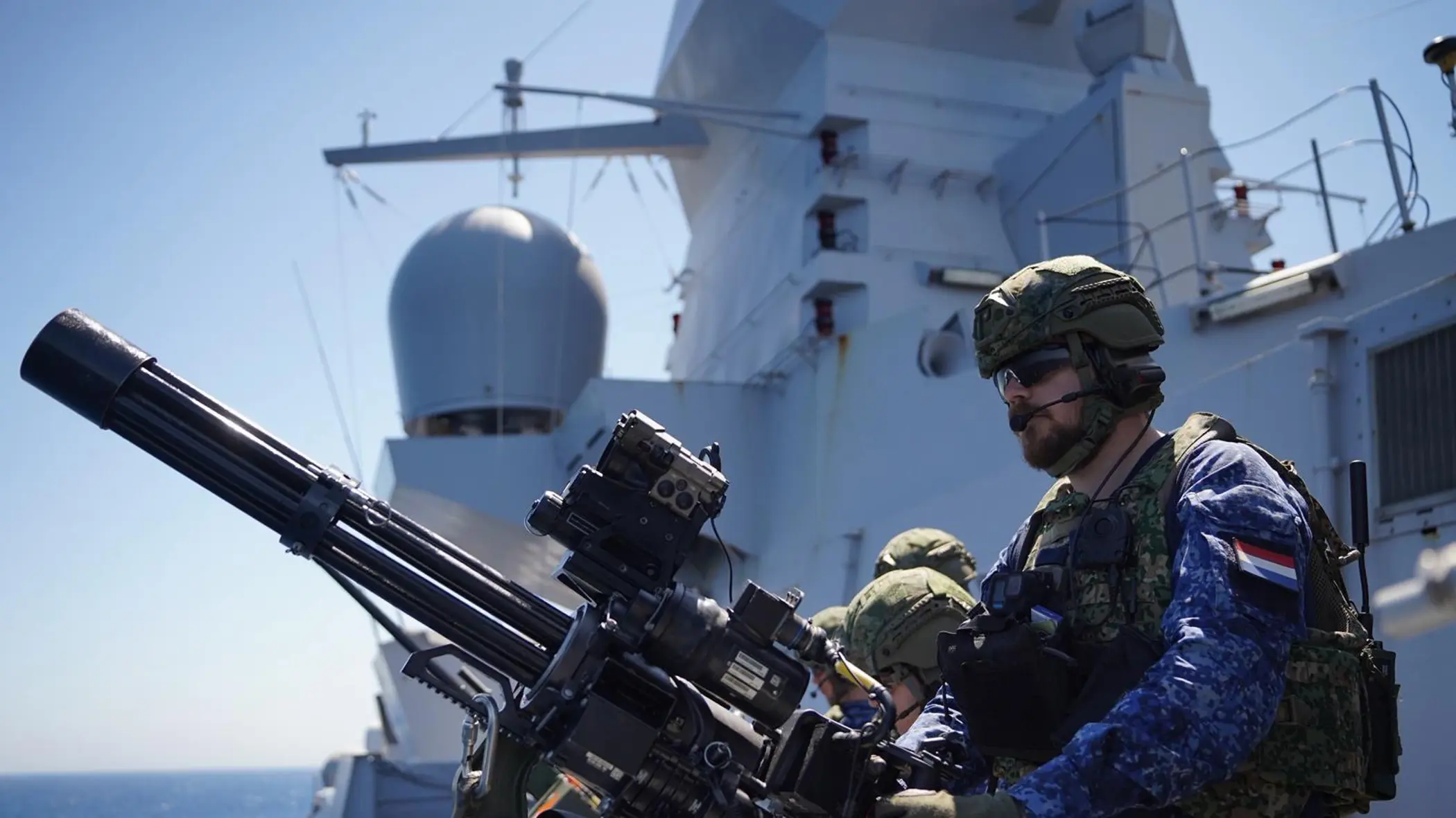 Een bemanningslid met baard staat achter een minigun op een van de dekken van Zr.Ms. De Ruyter