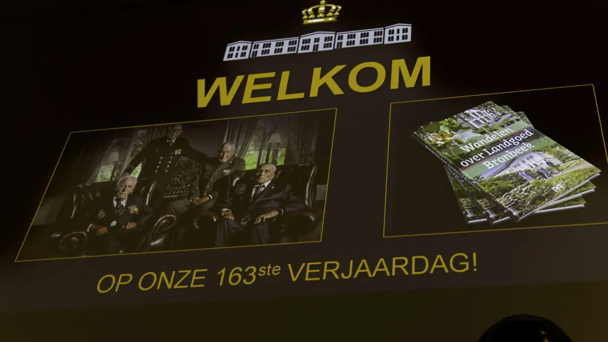 Een groot scherm met daarop een foto van vier  veteranen en een beeld van de wandelgids. Qua tekst staat er: welkom op onze 163e verjaardag. 
