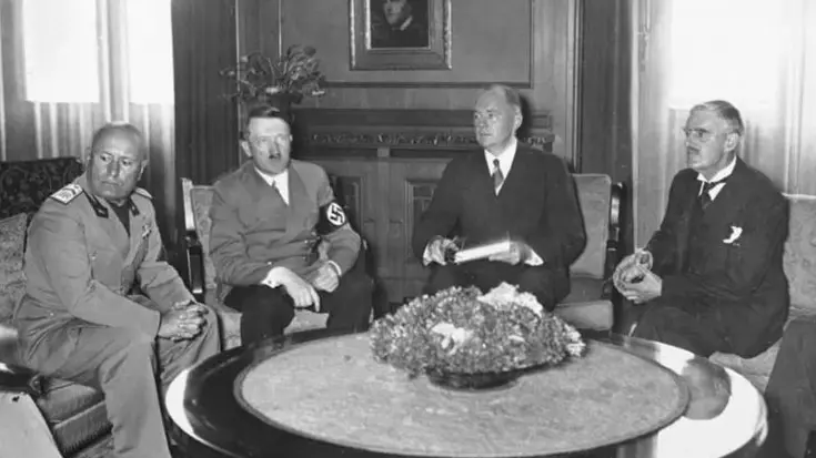 Vier mannen in pak zitten rond een ronde tafel met bloemstuk in een klassieke kamer met portret aan de muur.
