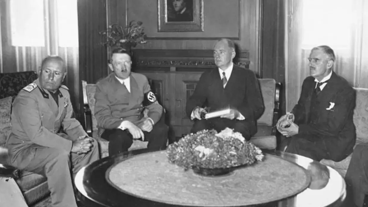 Vier mannen in pak zitten rond een ronde tafel met bloemstuk in een klassieke kamer met portret aan de muur.