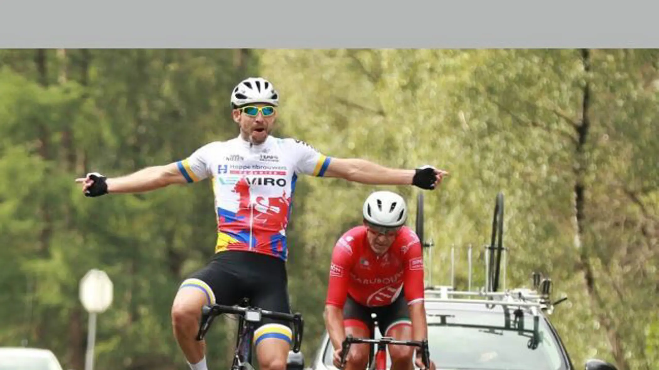 Twee wielrenners in wedstrijdtenue, waar de voorste juichend zijn armen spreidt, terwijl een auto met fietsen op het dak achter hen rijdt op een weg.