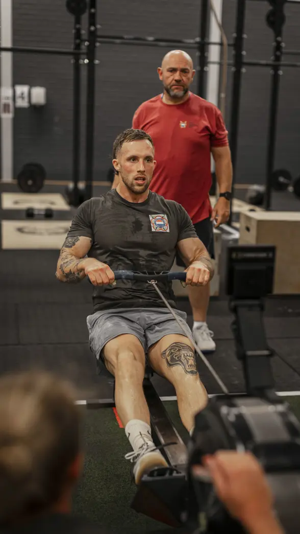 Een man traint op een roeimachine in een sportschool. Hij heeft tatoeages en draagt sportkleding. Een trainer kijkt toe. Apparaat staat centraal.