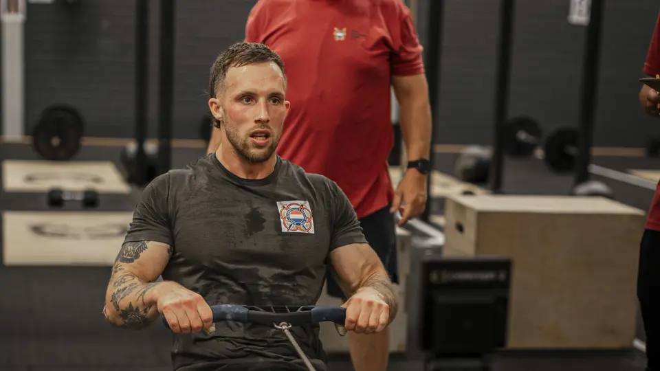 Een man traint op een roeimachine in een sportschool. Hij heeft tatoeages en draagt sportkleding. Een trainer kijkt toe. Apparaat staat centraal.