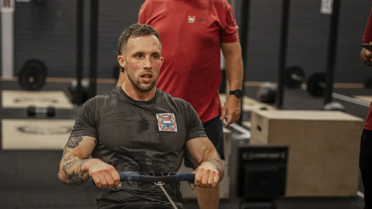 Een man traint op een roeimachine in een sportschool. Hij heeft tatoeages en draagt sportkleding. Een trainer kijkt toe. Apparaat staat centraal.
