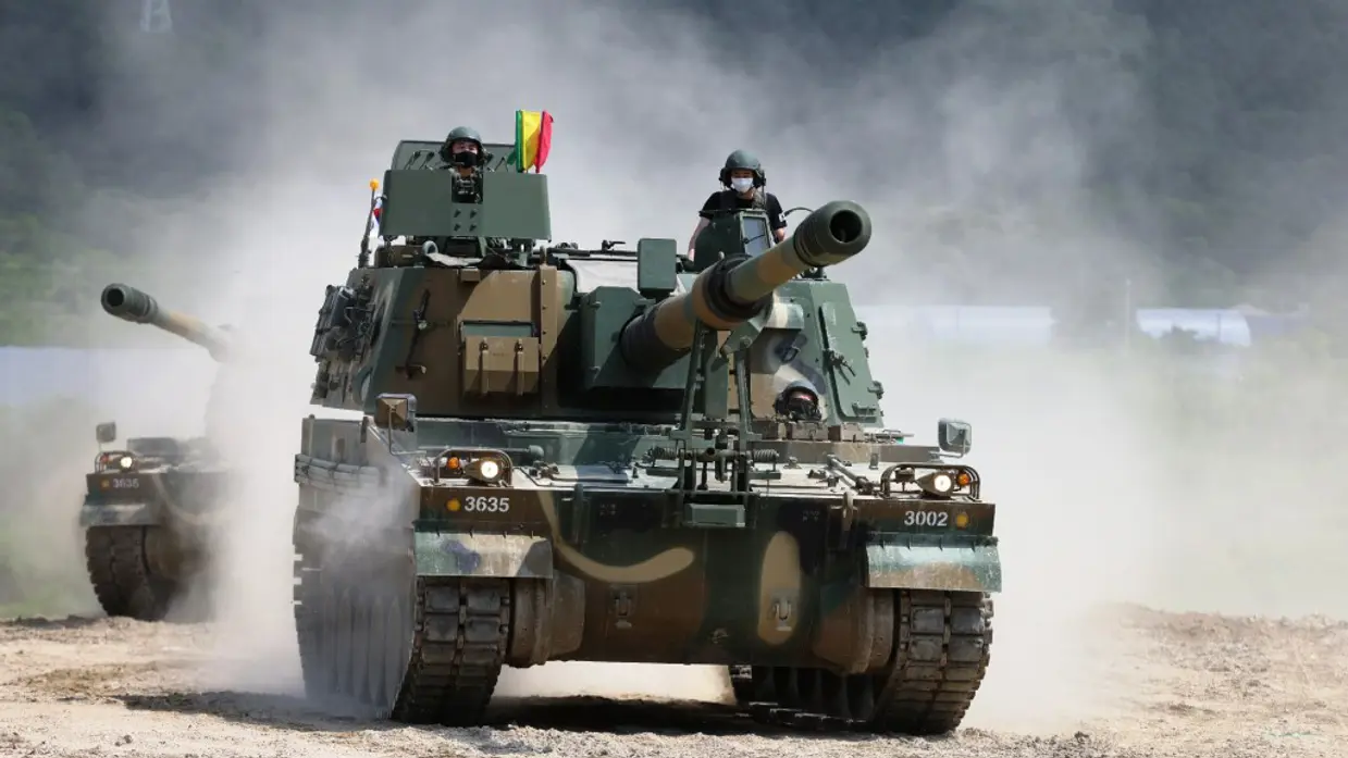 Een zwaar militair voertuig met een kanon rijdt over een stoffig terrein, terwijl een militair boven een luik uitsteekt.