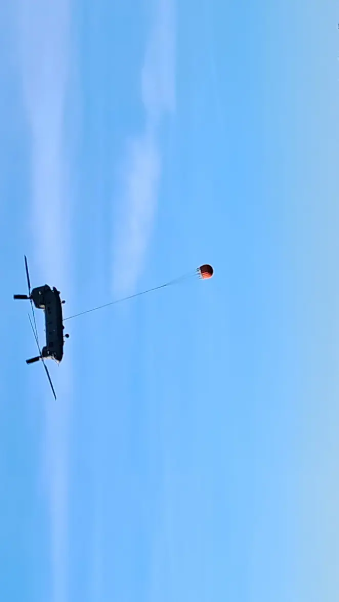 Een grote helikopter vliegt laag boven rustig, blauw water met een lange kabel en een felgekleurde vlag of bal eronder, kustlijn op de achtergrond. 
