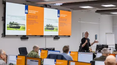 De facilitator van de CDF staat voor de groep en maakt gebruik van zijn handen om de situatie uit te leggen. De aanwezige burgers en militairen luisteren naar hem.