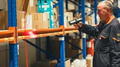  Een man staat voor een magazijnrek. In zijn handen heeft hij een handscanner, waarmee hij een barcode op het rek bliept.