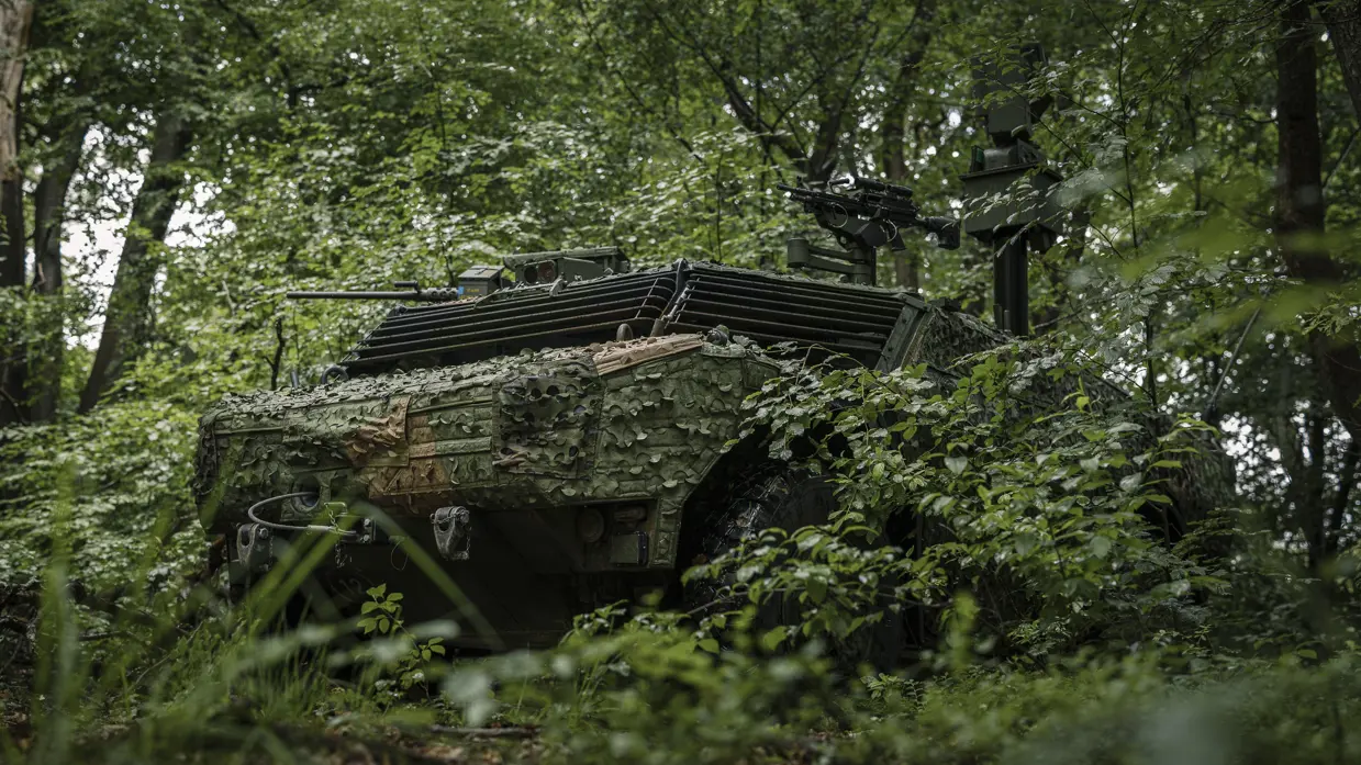 Een militair voertuig vol camouflage houdt zich schuil in een bos. 