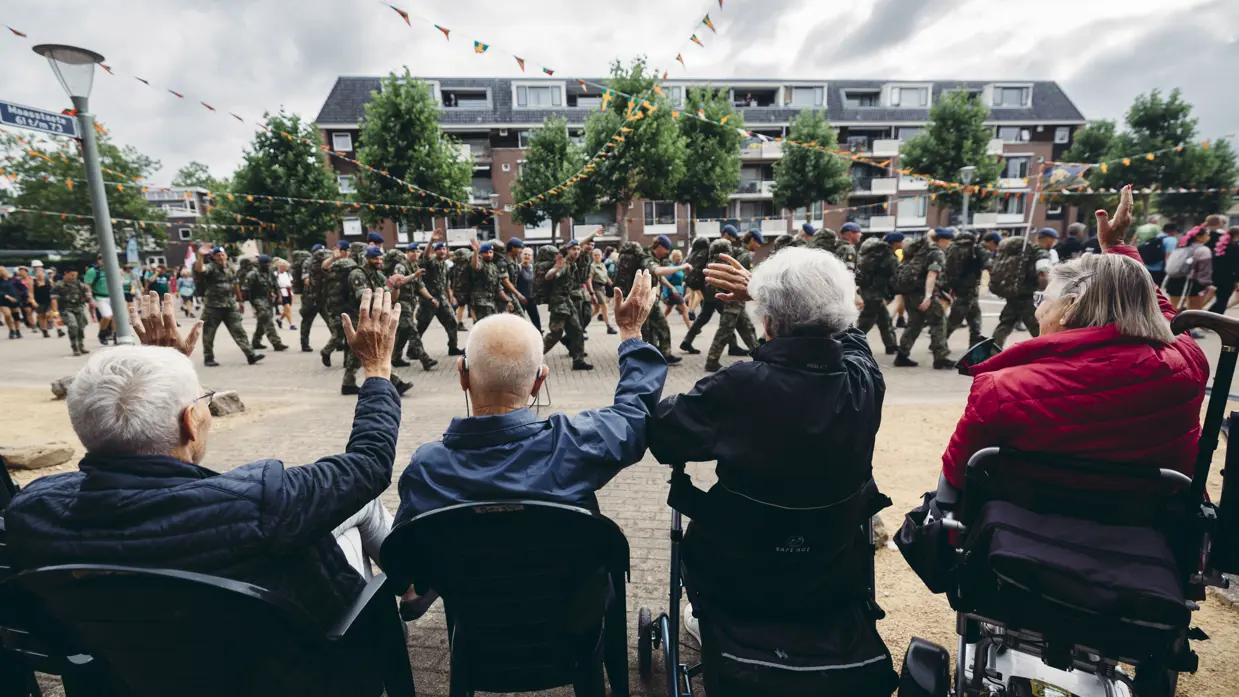 Een groepje oudere toeschouwers, op de rug gezien, moedigt een detachement van de Marechaussee aan tijdens de Nijmeegse 4Daagse. 