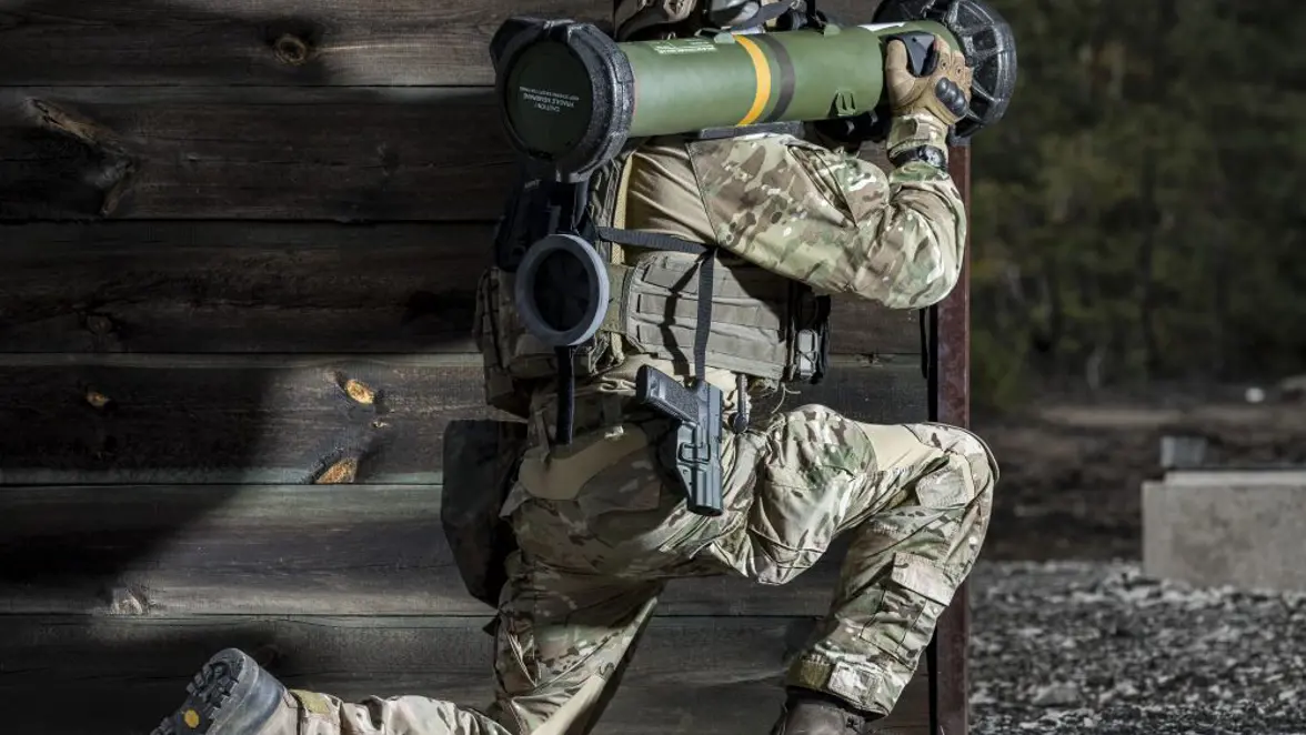 Een militair zit geknield achter een houten muurtje. Op zijn schouder rust het antitankwapen Spike SR.