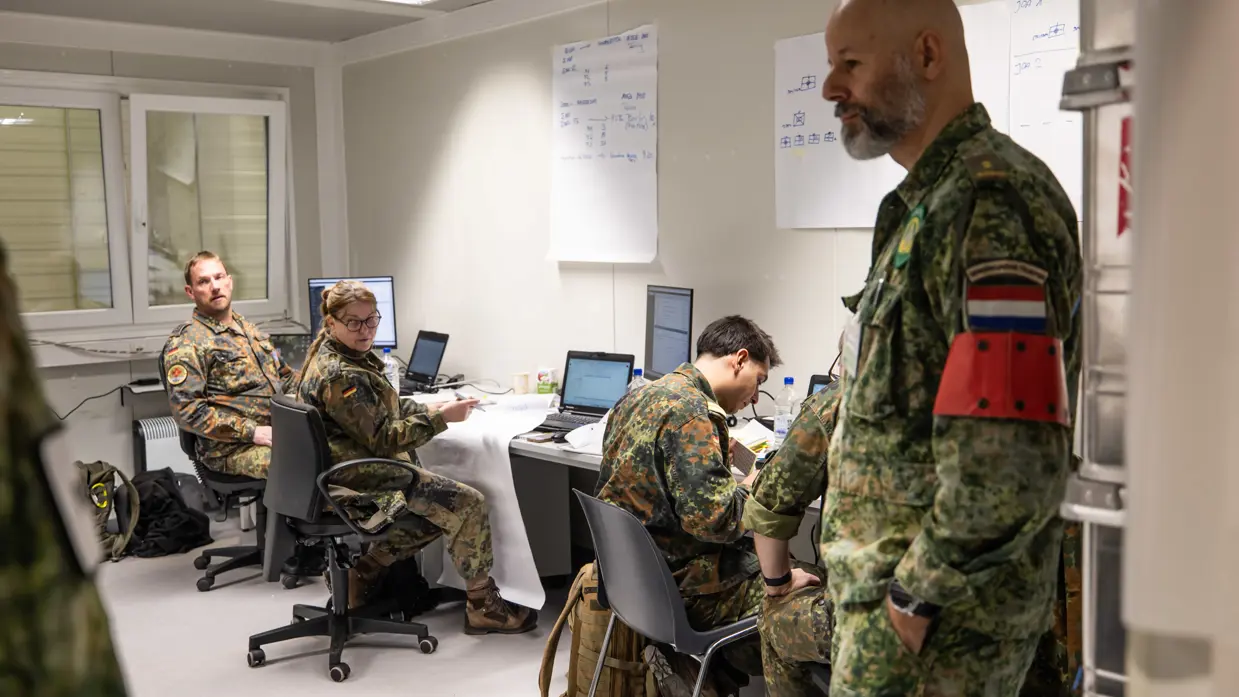 Links: Militairen in uniform werken in een kantoorruimte met laptops en papieren. Een man met Nederlandse vlag op zijn arm staat te luisteren. Kaarten hangen aan de muur. Rechts: Militairen in verschillende uniformen bespreken informatie achter een computer. Een vrouw met Italiaanse vlag op haar mouw kijkt toe, terwijl een man nadenkt.
