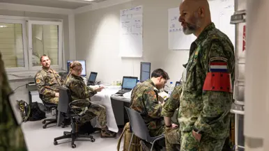 Links: Militairen in uniform werken in een kantoorruimte met laptops en papieren. Een man met Nederlandse vlag op zijn arm staat te luisteren. Kaarten hangen aan de muur. Rechts: Militairen in verschillende uniformen bespreken informatie achter een computer. Een vrouw met Italiaanse vlag op haar mouw kijkt toe, terwijl een man nadenkt.