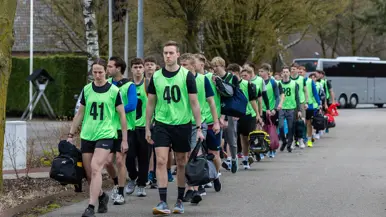 Een groep jongeren in felgroene hesjes met nummers staat in het gelid. Een militair in uniform geeft instructies op een parkeerterrein met bomen en voertuigen.