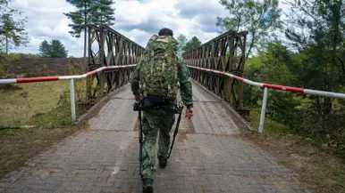 Een militair in camouflage met rugzak loopt over een houten brug met rode-witte afzetting in een bosrijk gebied.