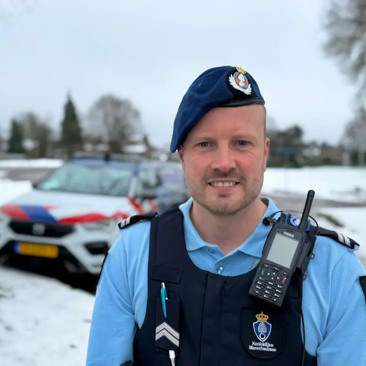 Portret van wachtmeester van de Marechaussee met voertuig op de achtergrond.